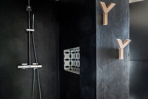 Un baño moderno con pared negra, ducha con detalles cromados y letras de madera formando la letra 'Y' en la pared.