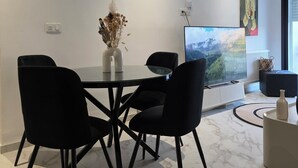 Ett modernt matområde med ett glasbord, svarta stolar, en vas med torkade blommor, en platt-TV och en tavla på väggen.