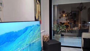 Ett modernt vardagsrum med en platt-TV, en svart pall, en flätad korg och ett stort fönster med utsikt över omgivningen.