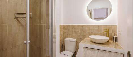 Et badeværelse med glasbrusekabine, toilet i hvid, rundt spejl og beige vask med guldfarvet armatur.