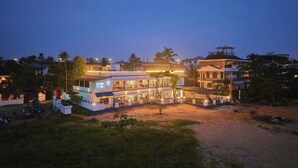 Ein mehrstöckiges Hotel mit überdachter Außenbestuhlung, umgeben von Grünpflanzen und Palmen.