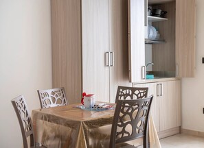 Ein Essbereich mit einem Tisch, gedeckt für vier Personen, umgeben von Stühlen mit aufwendigen Rückenlehnen. Im Hintergrund ist ein Schrank mit Glastür sichtbar, der Küchenutensilien beherbergt.