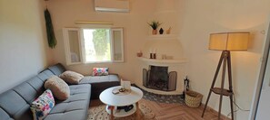 A living room with a sofa, a small table, a fireplace, a lamp, and a window. - Peaceful village stay (Karousades)