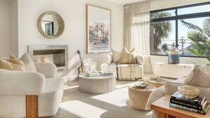 A modern living room with a fireplace, comfortable seating, a large window with curtains, and a view of palm trees. - Camps Bay Luxury Family Villa with Ocean Views (Cape Town)