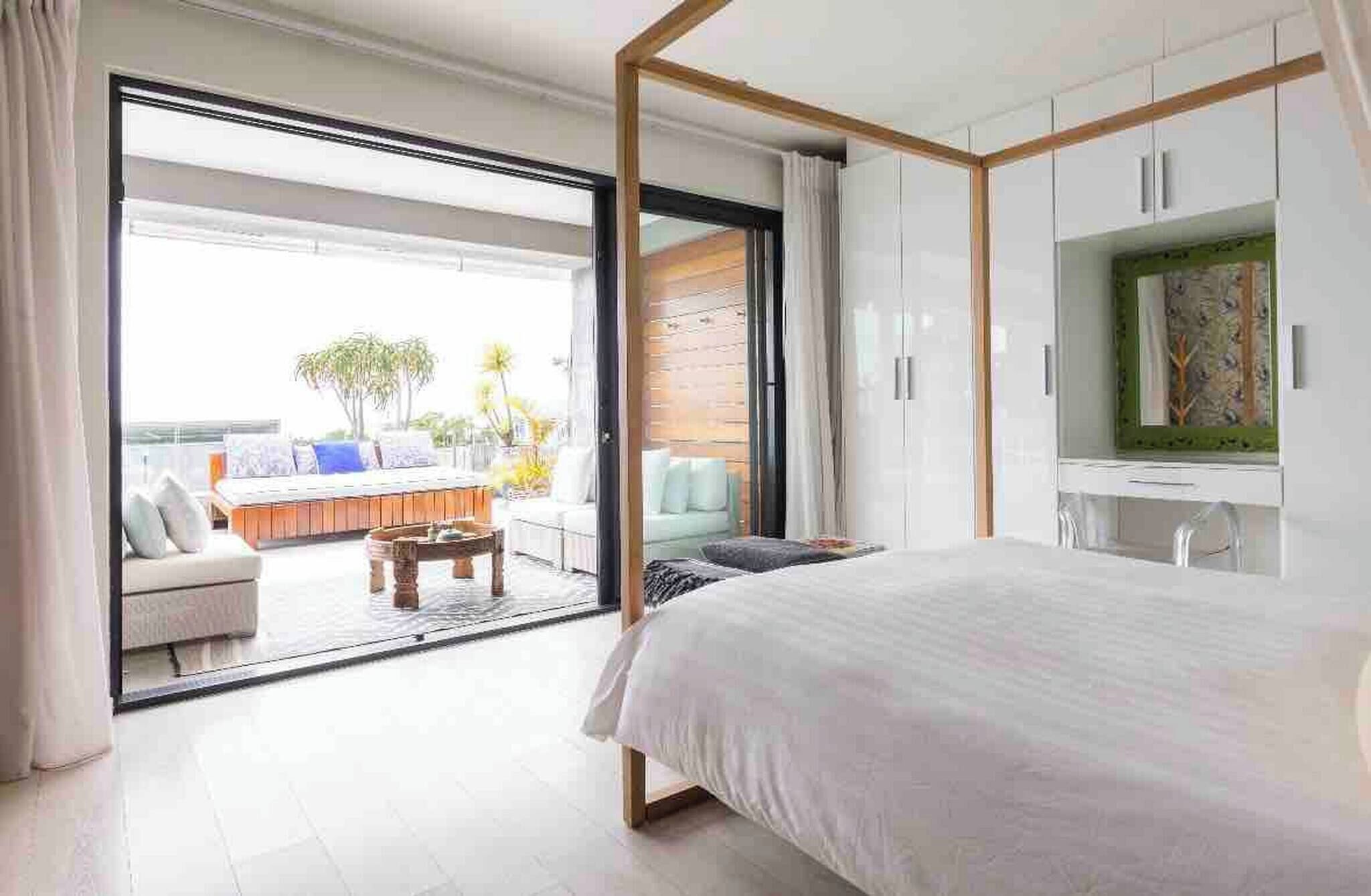A bedroom with a large bed, a TV, and a sliding glass door leading to a balcony with outdoor seating.