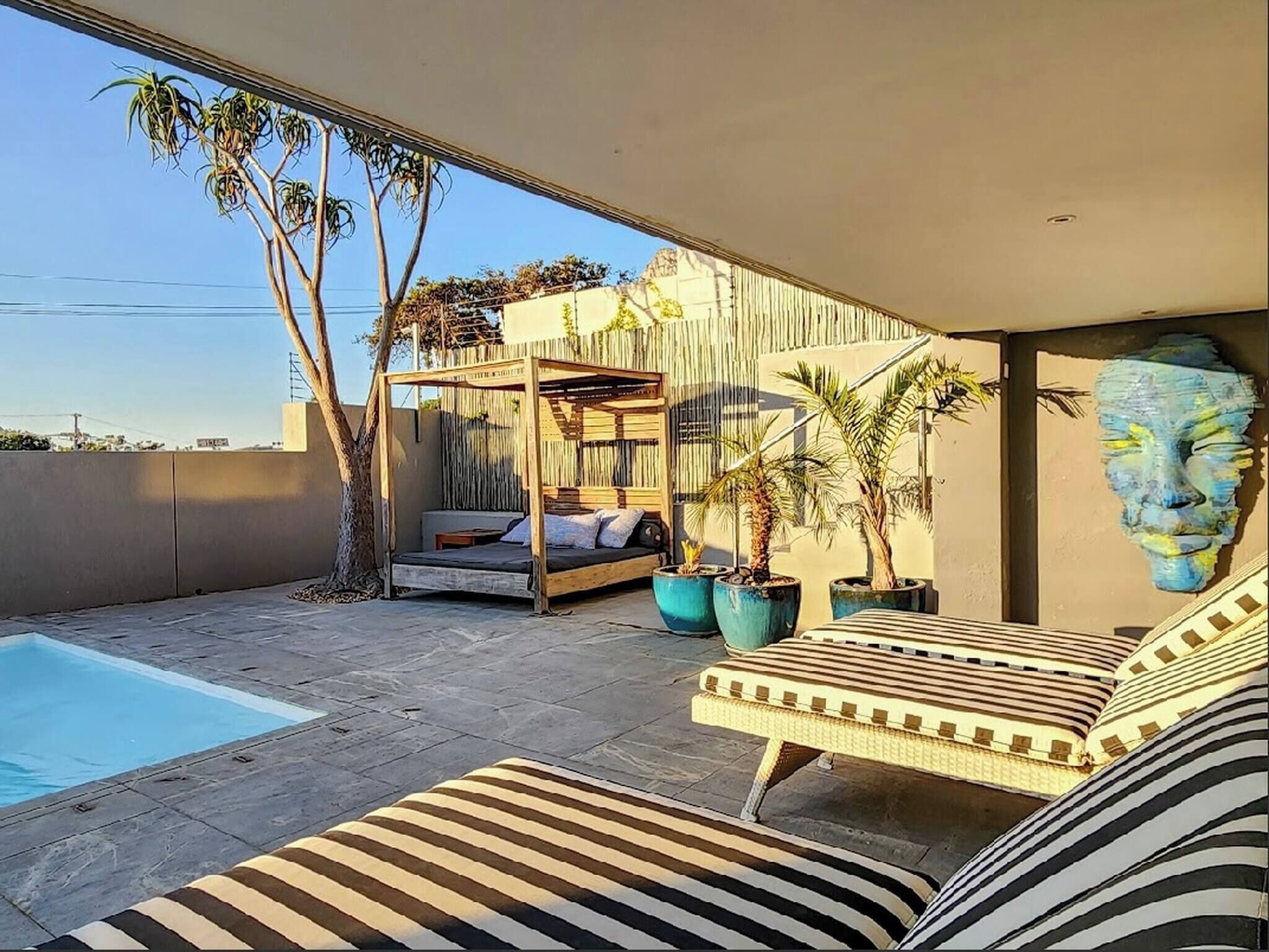 A modern outdoor living space with a pool, a wooden deck, a covered seating area, and potted plants.