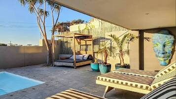 A modern outdoor living space with a pool, a wooden deck, a covered seating area, and potted plants.