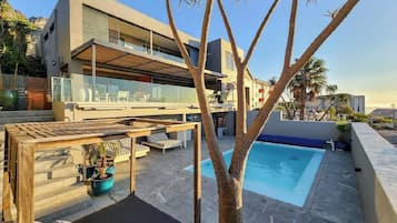 A modern two-story building with a rooftop pool, outdoor seating, and a tree in the foreground.