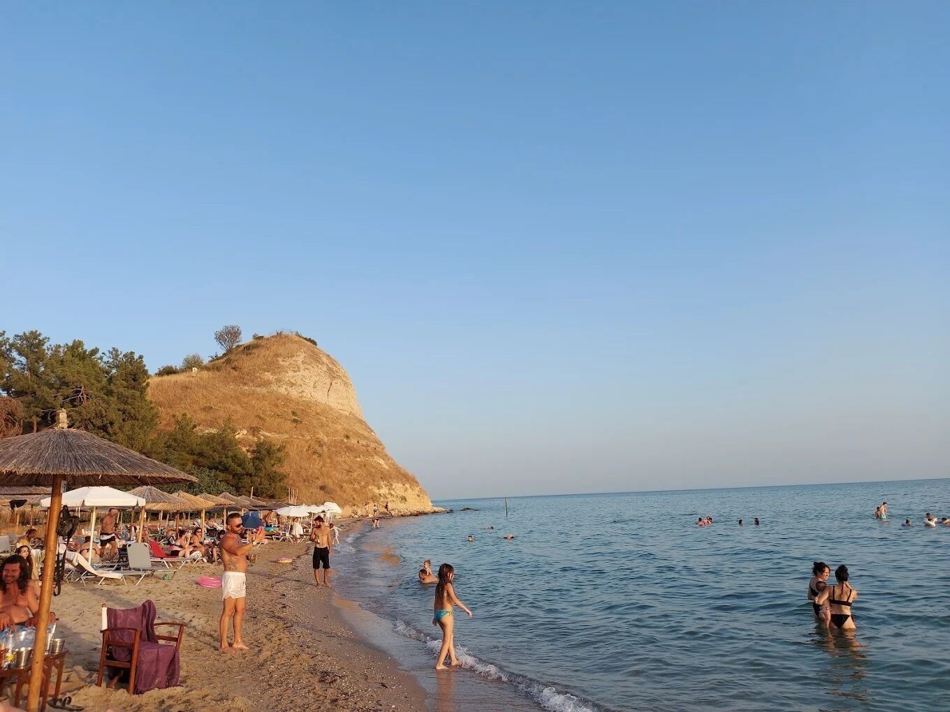 Ein Strand mit badenden und sonnenbadenden Personen, Sonnenschirmen und einer Hügelkette im Hintergrund.