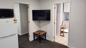 Une pièce de taille modeste, avec un réfrigérateur blanc, un téléviseur à écran plat fixé au mur, une petite table en bois et un meuble blanc avec une chaise.
