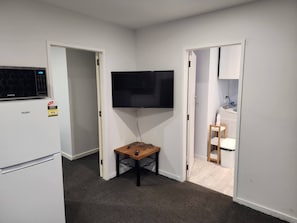 A compact room with a white refrigerator, a flat-screen TV mounted on the wall, a small wooden table, and a white cabinet with a chair.
