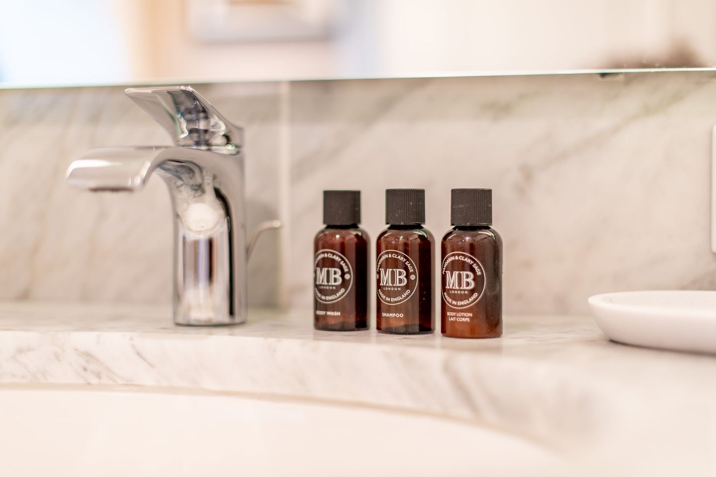 A bathroom with a marble countertop, a glass shower enclosure, and three bottles of MB London products.
