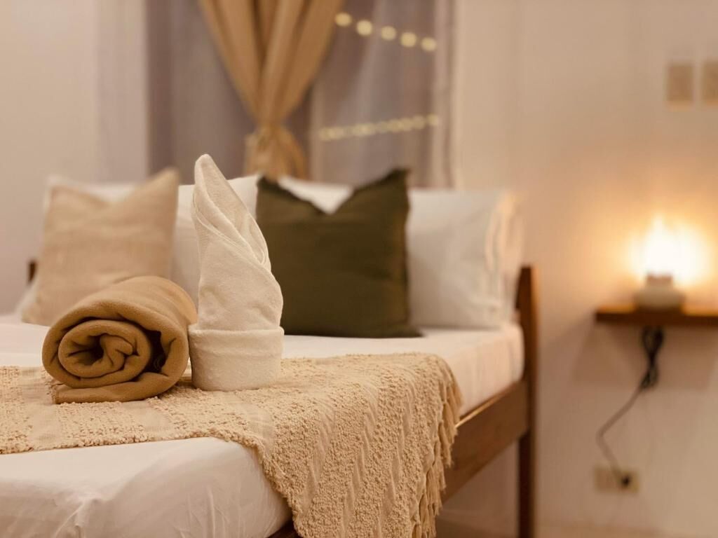 A neatly made bed with rolled towels and a blanket, a pillow, and a bedside table with a lamp.