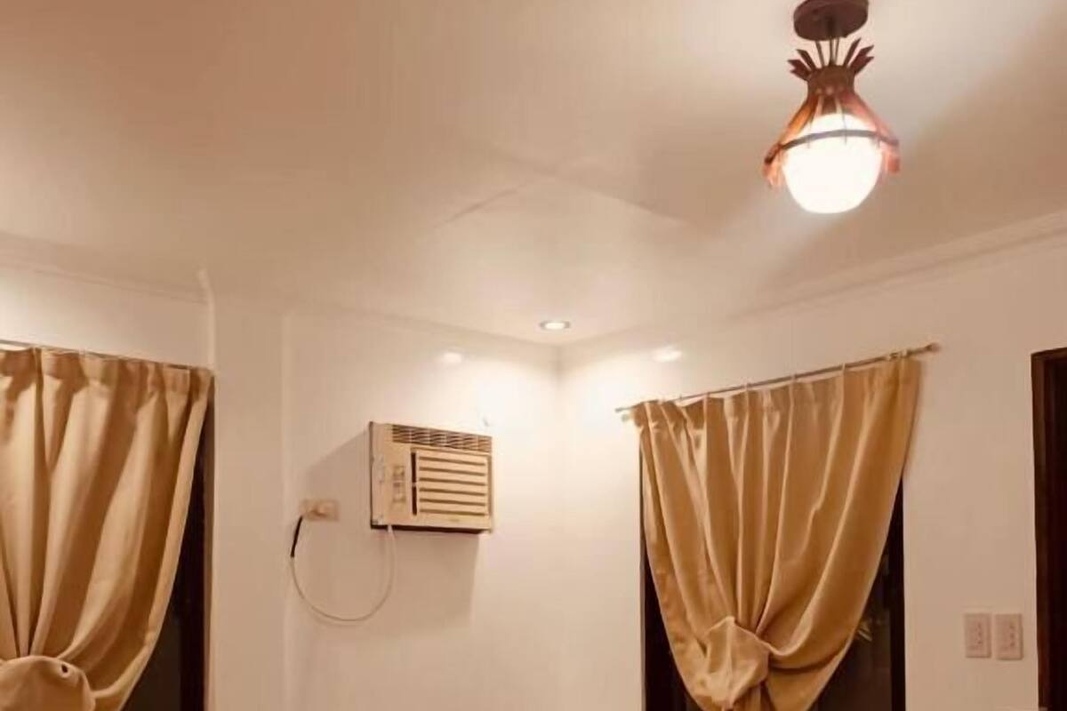 A hotel room with a bed, curtains, an air conditioning unit, and a ceiling light fixture.