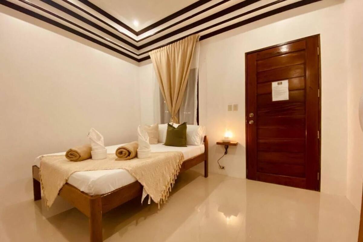 A bedroom with a bed, pillows, a bedside table, a wall-mounted lamp, a door, and a window with curtains.