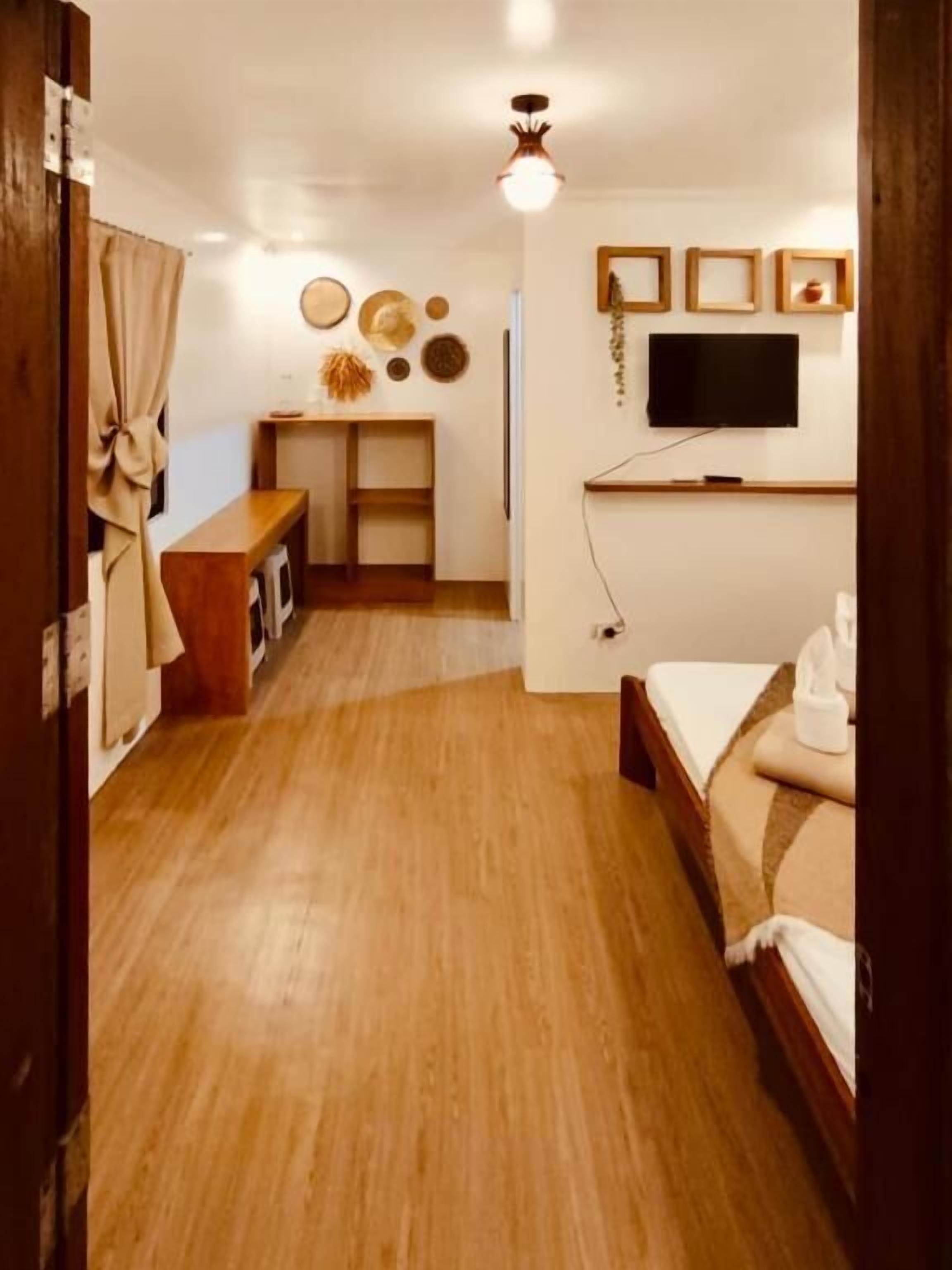 A compact hotel room with a wooden floor, a bed, a small desk, and a television mounted on the wall.