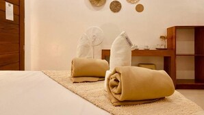 Une chambre d’hôtel avec un lit, deux serviettes, un ventilateur et une petite armoire en bois.