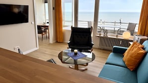 Quarto de hotel moderno com uma janela grande com vista para o mar, uma TV de tela plana na parede, uma mesa de centro redonda de vidro, um sofá azul e uma área de jantar com mesa e cadeiras.