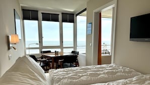Quarto de hotel com cama, mesa com cadeiras, televisão e vista para o mar.