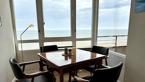 Uma área de refeições com uma mesa de madeira e quatro cadeiras, janelas amplas com vista para a praia e um abajur à esquerda.