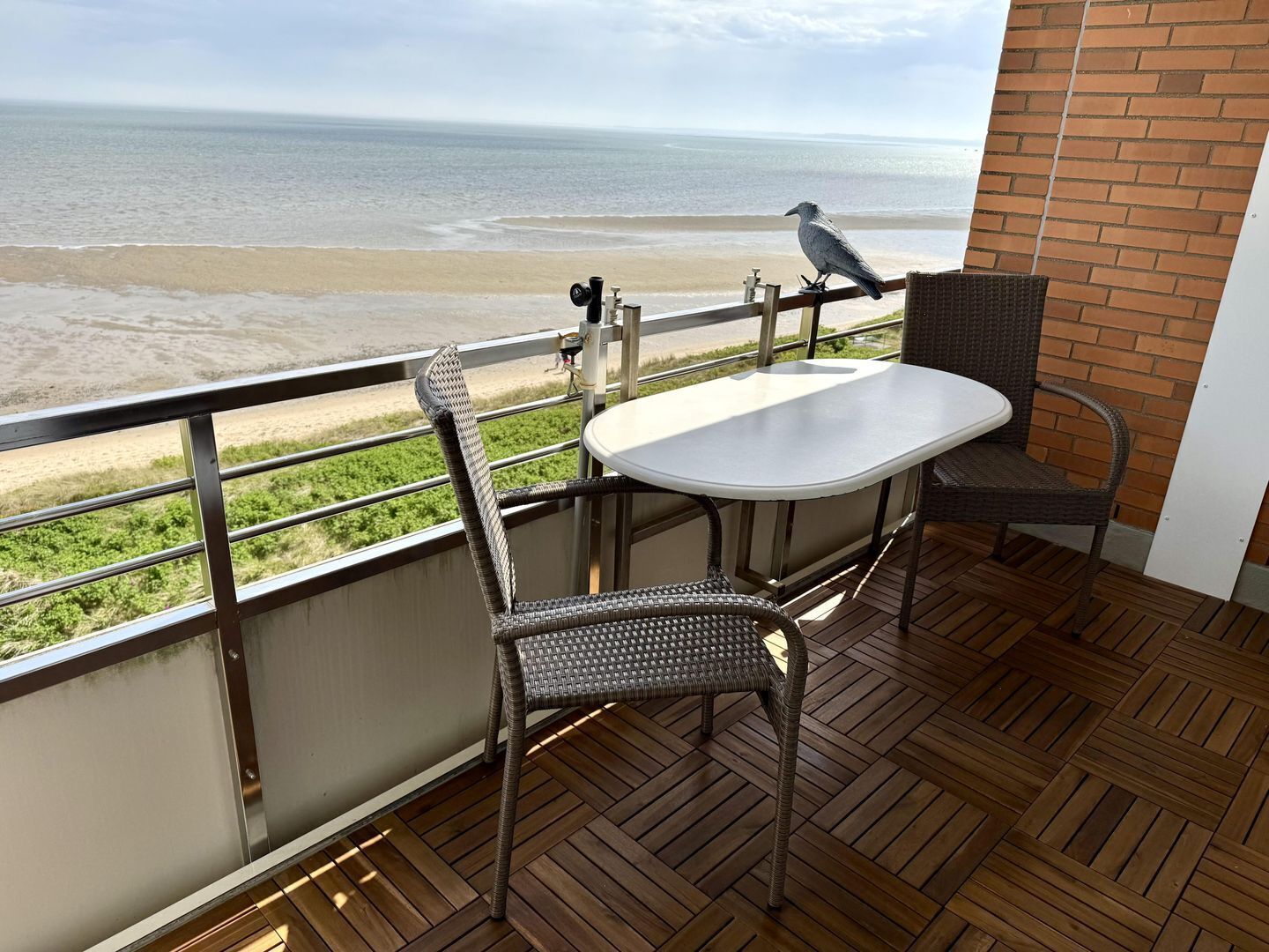 Un balcon donnant sur la plage, une table, deux chaises en osier et un oiseau perché sur la rambarde.