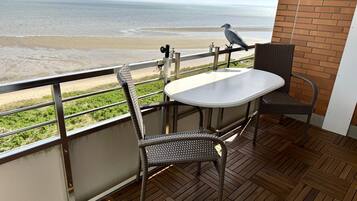A balcony with a view of the beach, a table, two wicker chairs, and a bird perched on the railing.