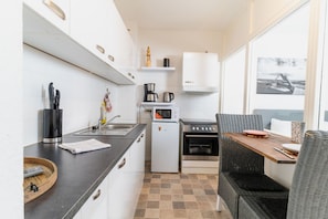 A compact kitchen with a sink, stove, and microwave, a dining area with a table and chairs, and a view through a window. - fewo1846 - Koje Sansibar - fewo1846 - Koje Sansibar / centrally located (Flensburg)