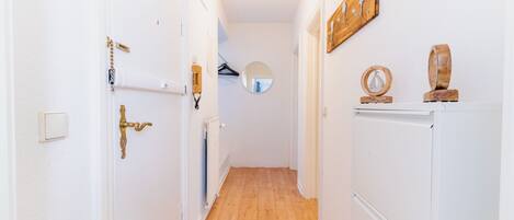 A narrow hallway with white walls, wooden flooring, a door with a brass handle, a coat rack, a mirror, and a radiator.