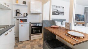 A compact kitchen with a microwave, coffee maker, and oven, a dining area with a table and chairs, and a large window with a view of the sea. - fewo1846 - Koje Sansibar - fewo1846 - Koje Sansibar / centrally located (Flensburg)