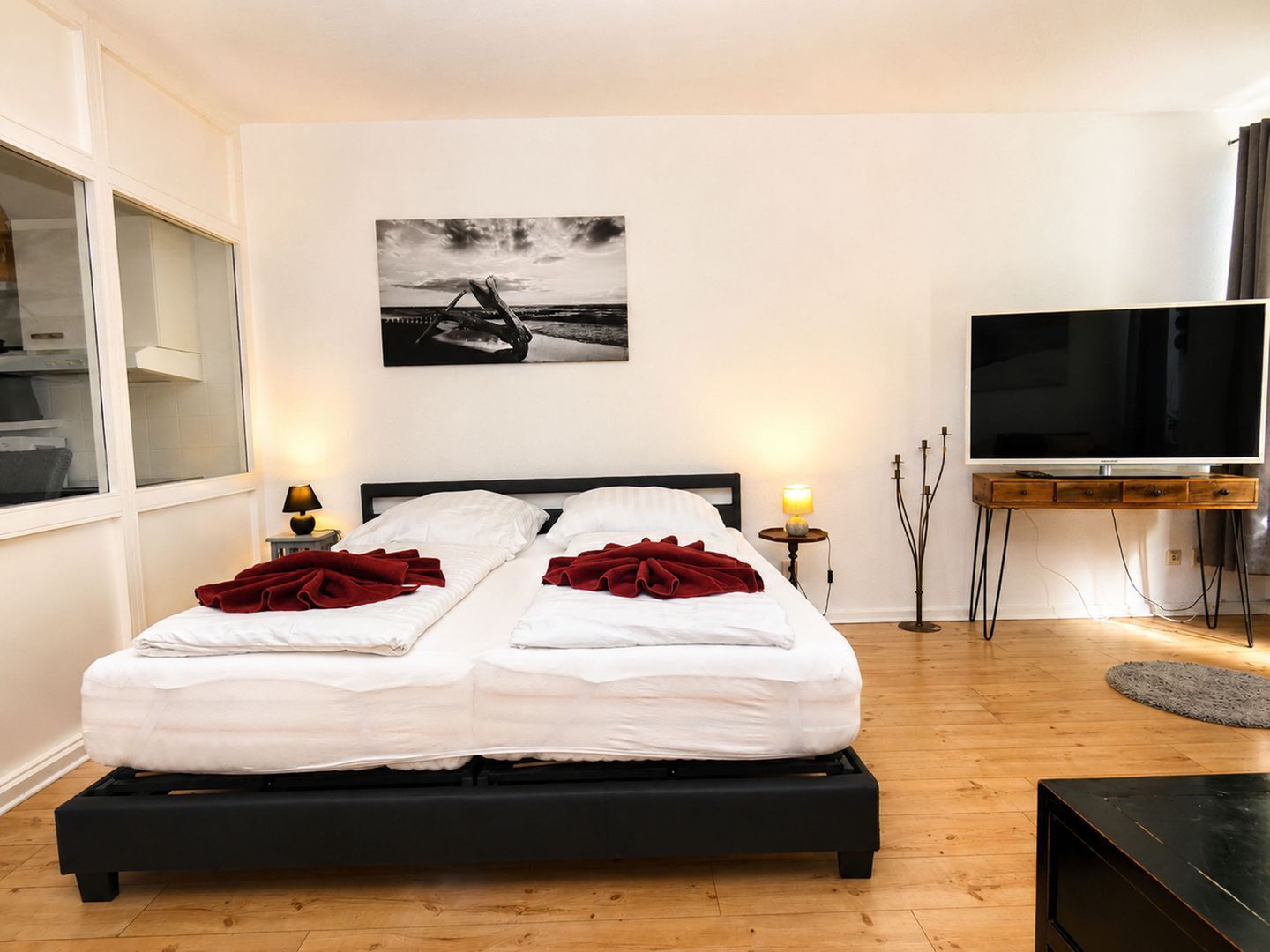 Una cama doble con sábanas blancas y almohadones rojos, una mesita de noche con una lámpara, un televisor sobre un mueble bajo y un cuadro en la pared.