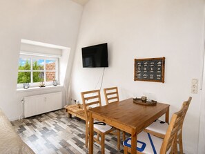Uma área de refeições com mesa e cadeiras de madeira, televisão fixada na parede e janela com vista para o telhado de um edifício.