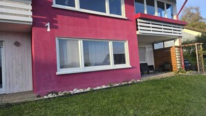 Une maison à deux étages, avec une façade rouge, des encadrements de fenêtres blancs et un petit jardin comprenant une pelouse et une terrasse en bois.