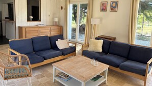 A living room with two dark blue sofas, a wooden coffee table, a wicker chair, and a view of a garden through large windows.