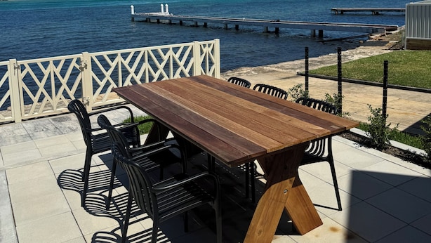 Un patio al aire libre con una mesa y sillas de madera, con vista a una masa de agua con un muelle y tierra a lo lejos.