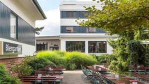 Ein Sitzbereich im Freien mit Tischen und Stühlen, ein Gebäude mit der Aufschrift 'Biergarten' und üppigem Grün.