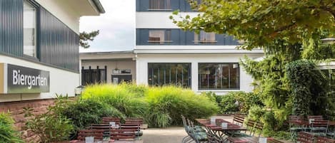 An outdoor seating area with tables and chairs, surrounded by greenery and a building with a sign reading 'Biergarten'.
