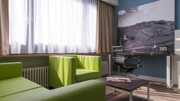 Habitación de hotel con sofá verde, un escritorio con silla, un radiador y un cuadro de paisaje grande en la pared.