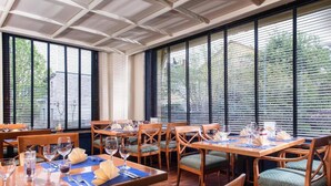 A dining area with tables set for four, wooden chairs with green cushions, and large windows with blinds. - Offenburg City by Belvilla-Family Room (Offenburg)