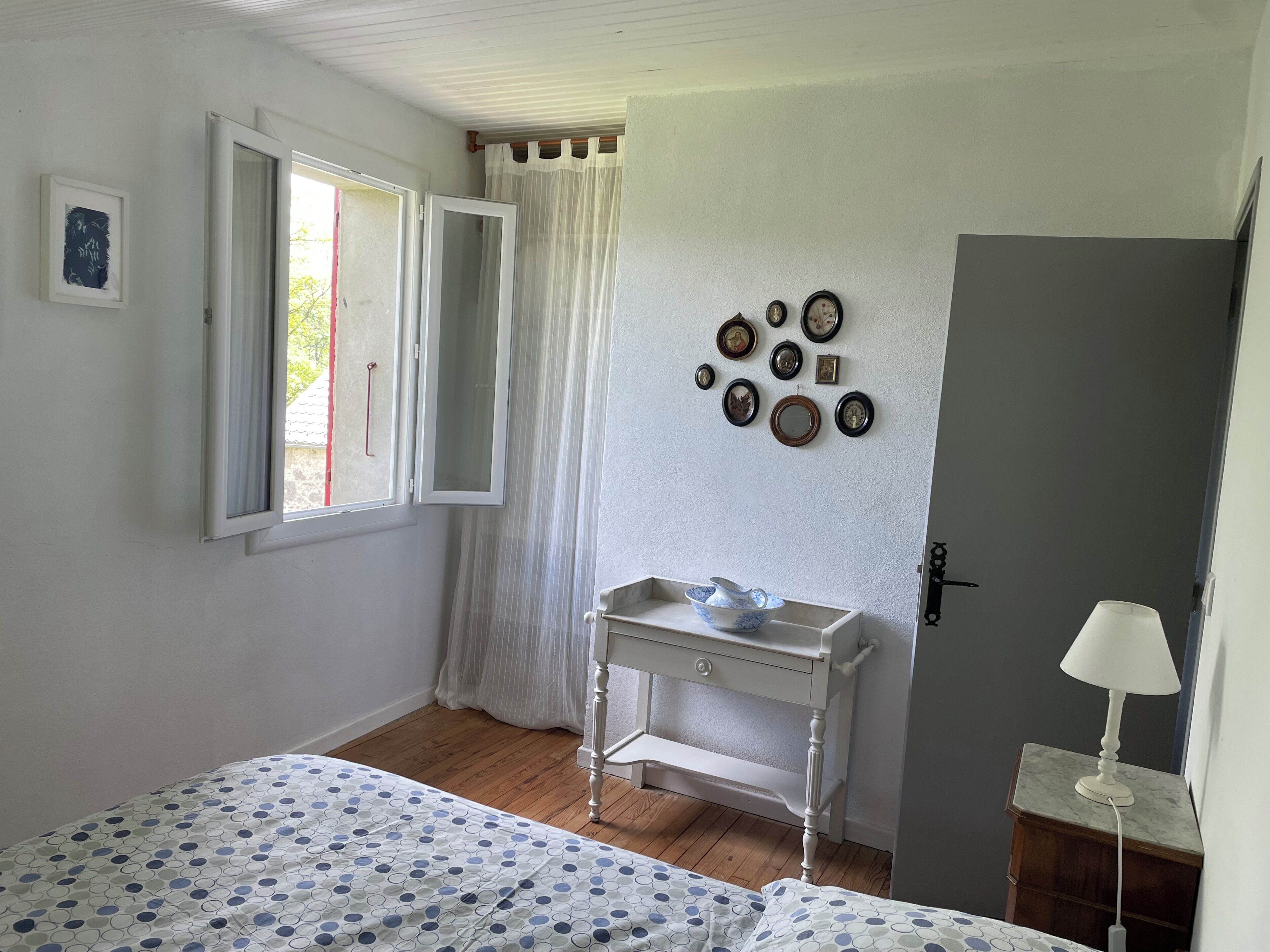 A bedroom with a bed, a bedside table, a lamp, a window with curtains, a door, and wall-mounted pictures.