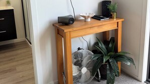 Una habitación con televisor montado en la pared, una mesa consola de madera con un altavoz, un ventilador y una planta en maceta.