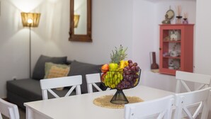 Een eettafel met een fruitschaal, witte stoelen, een grijze bank, een spiegel en een roze kast in een kamer.