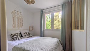 A bedroom with a bed, a window with curtains, a wall-mounted light fixture, and a wall decoration. - Appartement proche plage (Saint-Palais-sur-Mer)