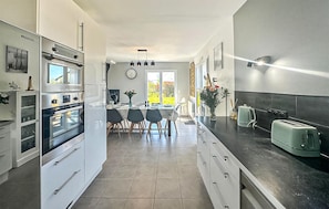 A modern kitchen with stainless steel appliances, a dining area with a table and chairs, and a view of the outdoors. - Charmante maison proche plage (Le Vivier-sur-Mer)