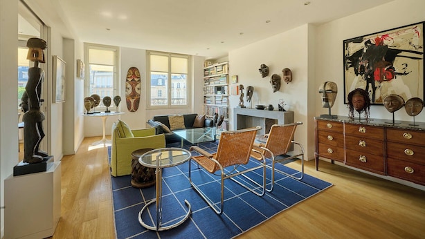 Een moderne woonkamer met een glazen salontafel, rieten stoelen, een open haard en een boekenkast vol boeken en decoratieve objecten.