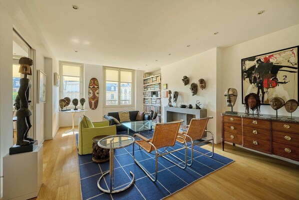 A modern living room with a glass coffee table, wicker chairs, a fireplace, and a bookshelf filled with books and decorative items. - DAUPHINE SQ City island - Elegant Apartment - 1 Bedroom - 1 Bath - Sleeps 2 (Paris)