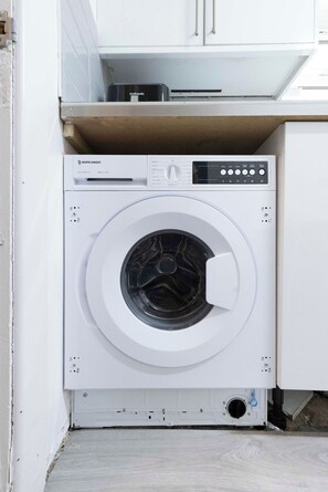 A front-loading white washing machine with a control panel, located in a kitchen area with white cabinets. - Spacious 4BR Home in Mile End – Amazing Location (Greater London)