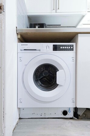 Un appareil compact, blanc, combinant une machine à laver et un sèche-linge à chargement frontal, doté d’un panneau de commande, et placé dans un coin d’une pièce dont les meubles de rangement sont également blancs.