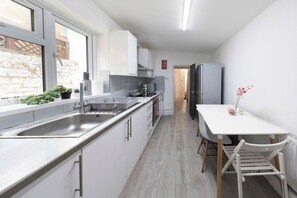 A modern kitchen with a sink, stove, and refrigerator. There is a window with plants on the counter, and a white table with chairs. - Spacious 4BR Home in Mile End – Amazing Location (Greater London)