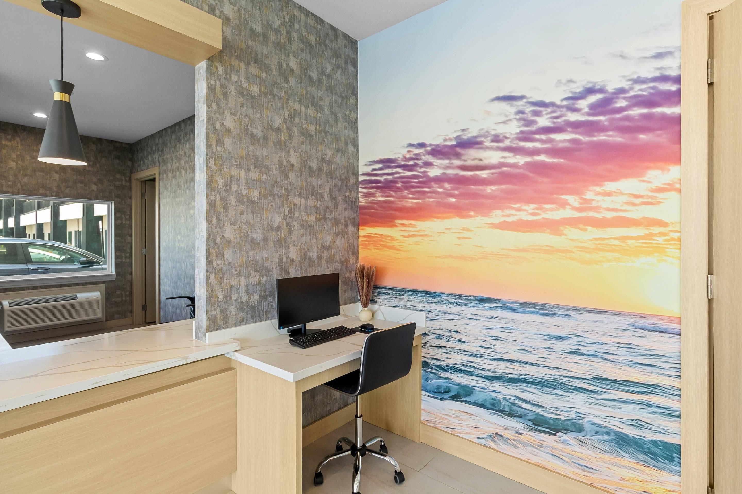 A modern office space with a desk, computer, and a large wall mural of a sunset over the ocean.