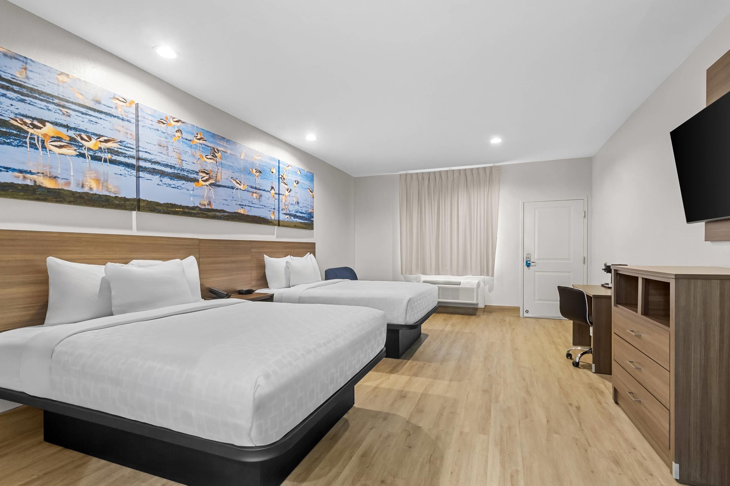 A hotel room with two beds, a desk, a chair, a TV, and a painting on the wall.
